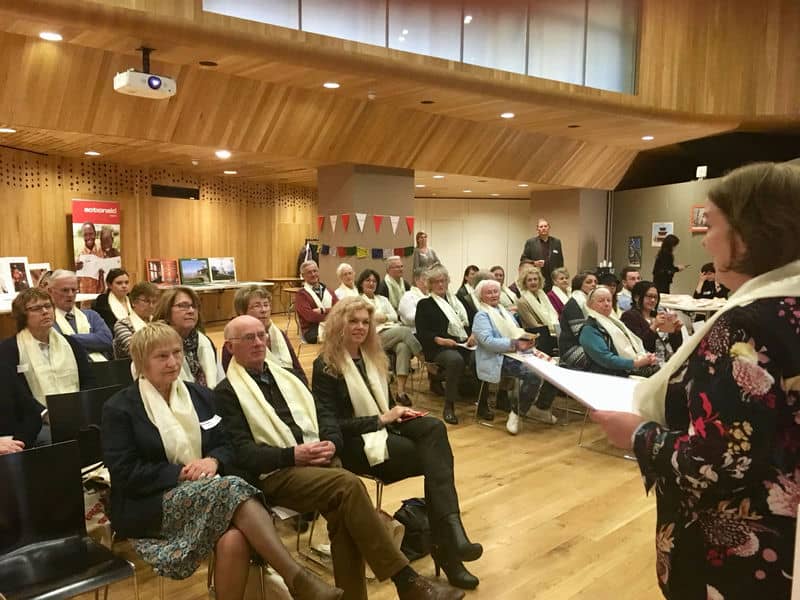 Audience at a seminar in a wooden hall, attendees wearing white scarves and seated while listening to a speaker.