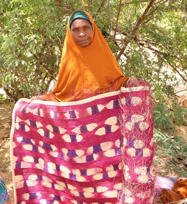 The image shows a woman standing outdoors, holding a brightly colored mat or textile. She is wearing an orange headscarf with a green undercap and is standing in front of a backdrop of leafy green trees. The textile she is holding features a striking pattern with red, purple, and cream colors.