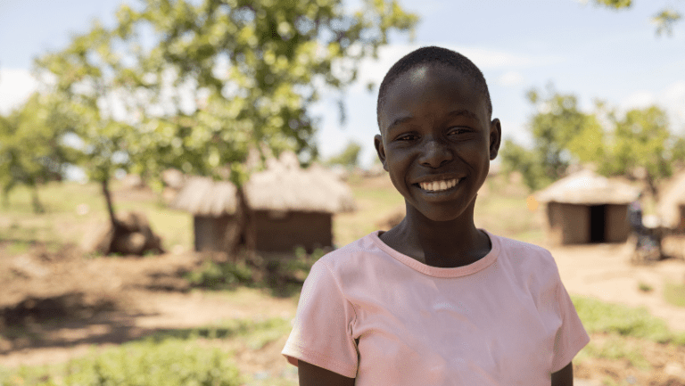 Charity, a smiling girl in a light pink t-shirt, stands outdoors in front of traditional huts and trees in her village.