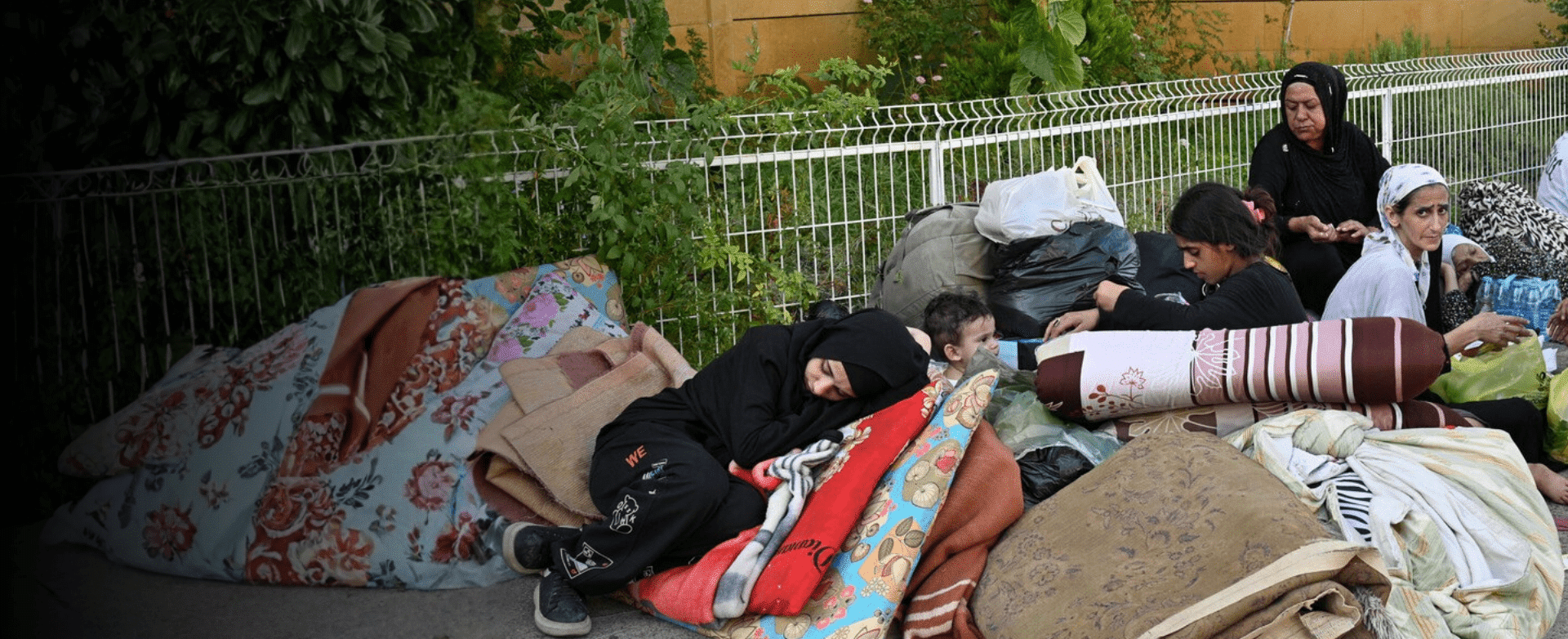 Women and children sit on the roadside with mattresses, bags and water bottles after being displaced by conflict in the Middle East.