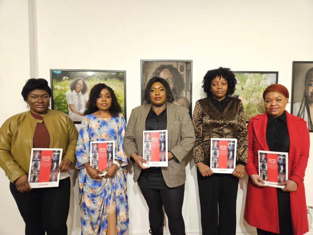 Five women holding pamphlets stand in front of portraits, showcasing a community event or workshop.