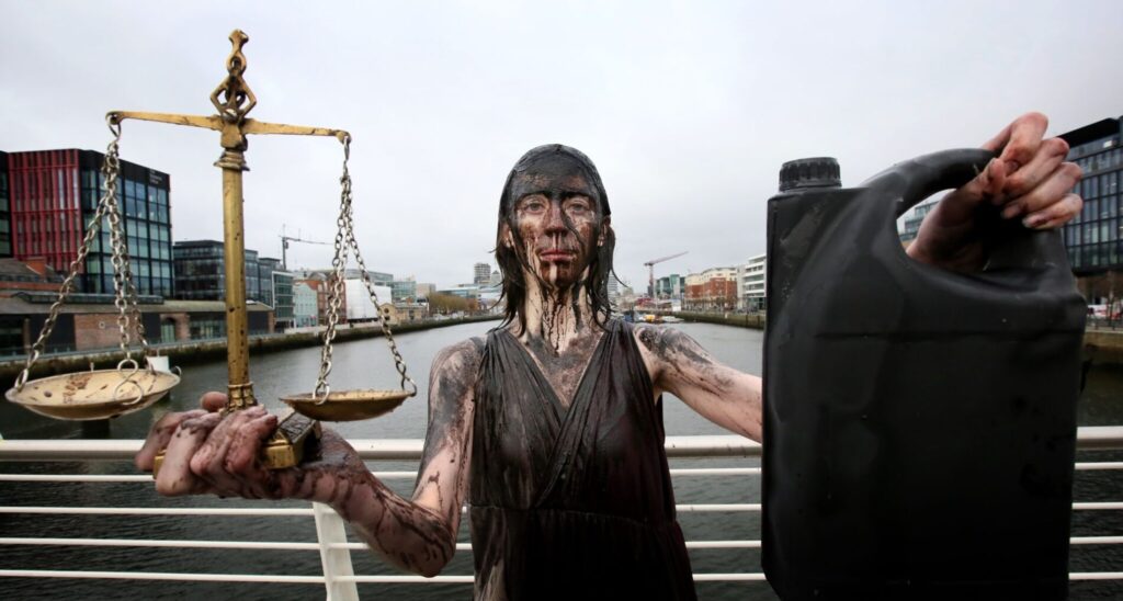 Protester covered in oil holds scales and a container, symbolizing environmental justice on a city bridge.