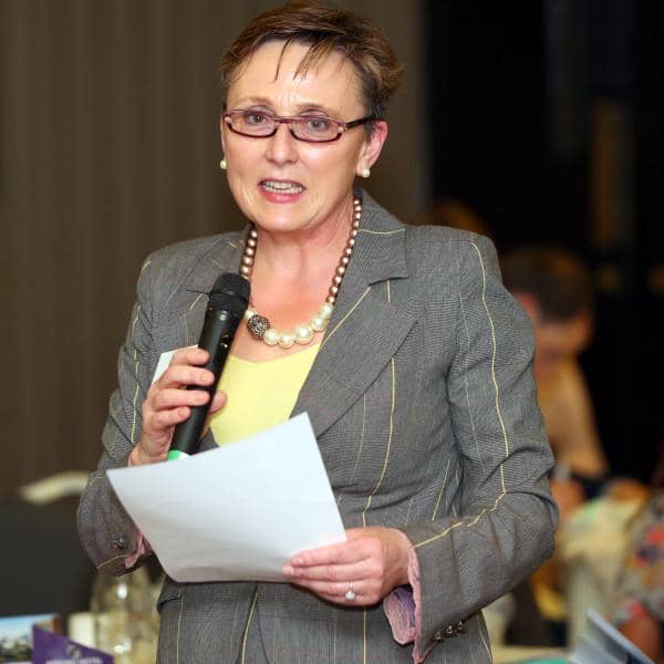 Woman delivering a speech with a microphone at a formal event, wearing a gray suit and glasses.