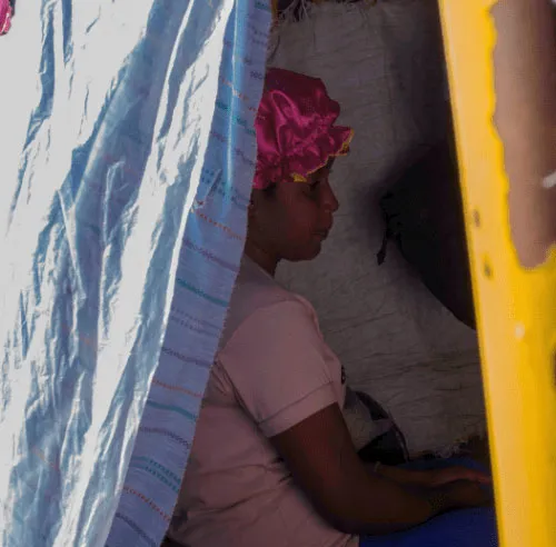Person wearing a pink bonnet sitting inside a tent, partially covered by a blue sheet.