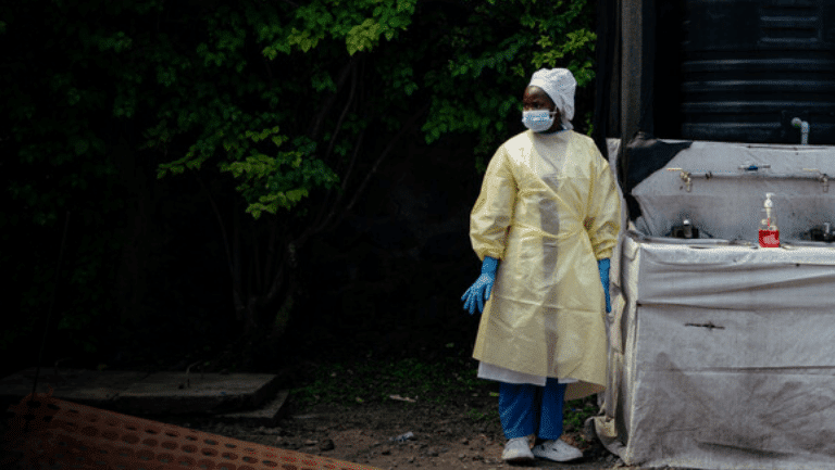 Portrait of a health worker wearing single-use protective clothing and a surgical cap at his checkpoint, where he oversees handwashing and temperature screening for all visitors and patients entering Kyeshero Hospital as part of Ebola prevention measures.