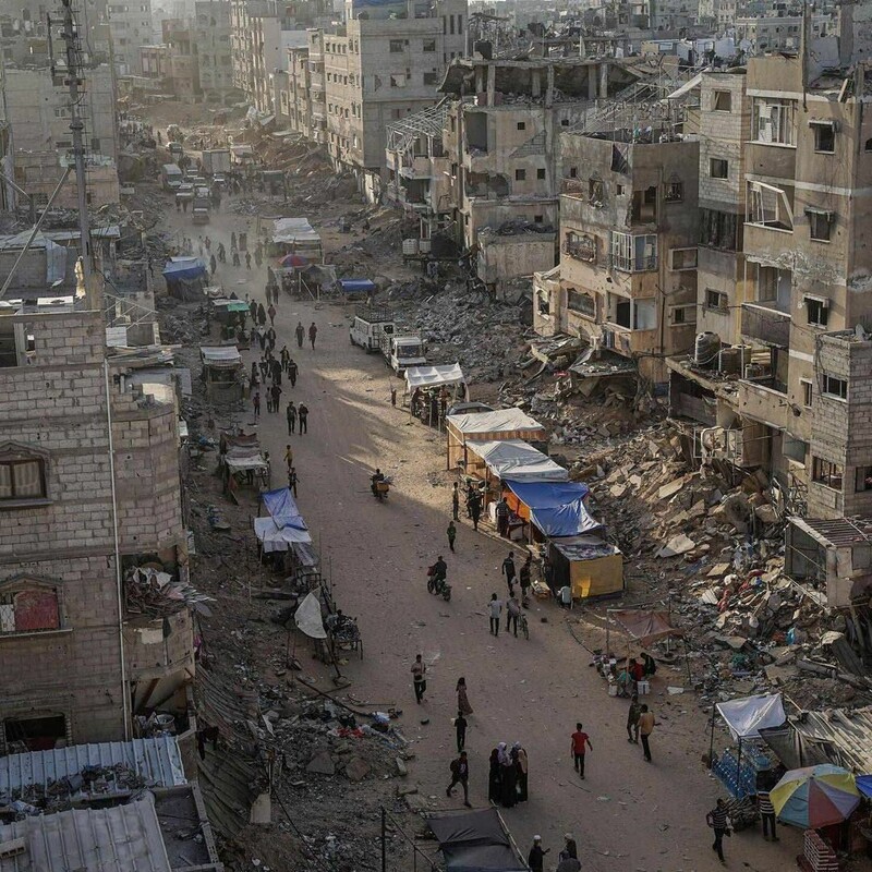 Aerial view of a city street with damaged buildings, rubble, and people walking past makeshift market stalls.
